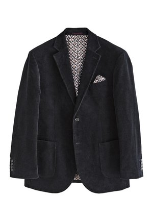 Mann in schwarzem Cordblazer mit Einstecktuch, dunklem Pullover, weißem Hemd, schwarzer Hose und schwarzen Schuhen, stehend und zur Seite schauend.