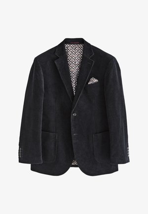 Mann in schwarzem Cordblazer mit Einstecktuch, dunklem Pullover, weißem Hemd, schwarzer Hose und schwarzen Schuhen, stehend und zur Seite schauend.