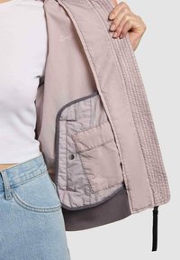 Person hält eine offene hellrosa Jacke, die Innentasche mit Druckknopf ist sichtbar, trägt ein weißes Oberteil und hellblaue Jeans.