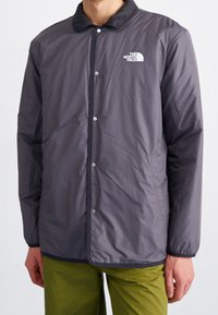 Szara ocieplana kurtka z zatrzaskami, czarnym kołnierzem oraz logo The North Face na lewej piersi. Posiada długie rękawy i prosty krój.