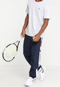 Šedé tričko s krátkým rukávem, tmavě modré sportovní kalhoty s bílými a červenými akcenty, držící tenisovou raketu. Bílé tenisky s černými detaily.