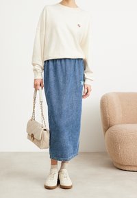 Sweatshirt crème clair, jupe maxi en denim bleu, sac à bandoulière beige et baskets blanches. Détails notables : un patch en forme de triangle sur la jupe.