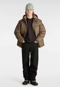 Kurtka puchowa w oliwkowej zieleni, z kapturem, elastycznymi mankietami i zapinana na zamek. Czarnie spodnie i jasnooliwkowa dziana czapka dopełniają stylizację.