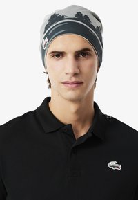 Berretto grigio chiaro con motivo a silhouette di alberi scuri, con una fascia nera e un logo sulla parte laterale, indossato sopra una polo nera.
