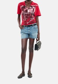 Polo rouge avec imprimé graphique, shorts en denim bleu clair, ceinture noire et chaussures plates noires. Les accessoires comprennent un petit sac à main noir.