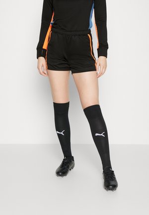 Person trägt schwarze Sportshorts mit orangefarbenen Streifen, schwarze kniehohe Puma-Socken und schwarze Fußballschuhe und steht auf einem hellen Boden.