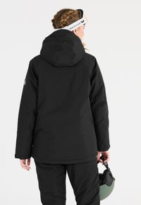 Schwarze wasserdichte Skijacke mit Kapuze, strukturiertem Stoff und einem Logo auf dem Ärmel. Das Modell hält einen grünen Skihelm.