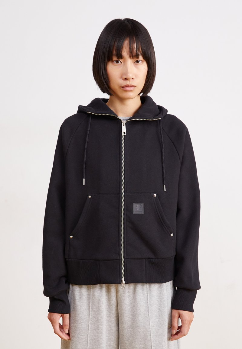 Sweat à capuche noir zippé en tissu doux, avec une capuche, deux poches avant et un patch logo. Accompagné d'un pantalon de survêtement gris.