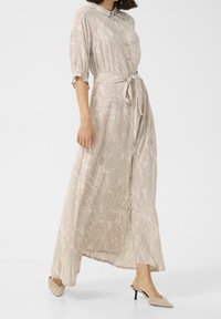 Robe midi beige clair avec un motif en vagues, manches courtes bouffantes, fermeture à boutons et détail de taille nouée. Portée avec des chaussures à talons hauts beiges.