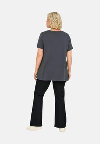 Grijze katoenen t-shirt met korte mouwen, gebogen zoom, gecombineerd met donker flare jeans en lichte schoenen. Model staat met de rug naar de camera.