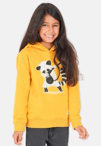 Gul huvtröja med en panda-grafik. Mjukt material, ribbad mudd och nederkant. Bärs över mörkgrå byxor.