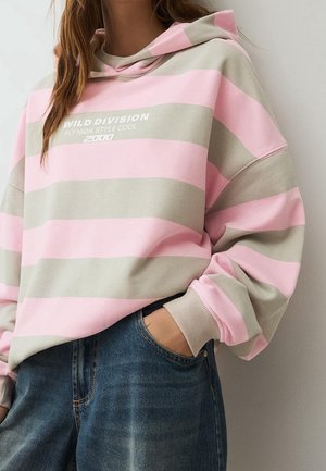 Person iført en pink og beige stribet hættetrøje med teksten "WILD DIVISION FLY HIGH, STYLE COOL 2000", kombineret med blå jeans, hånd i lommen.