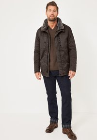 Braune Jacke mit grauem Kunstfellkragen, Knopfverschluss und Seitentaschen; getragen über einem braunen Pullover und dunklen Jeans mit Karostulpen, kombiniert mit braunen Stiefeln.