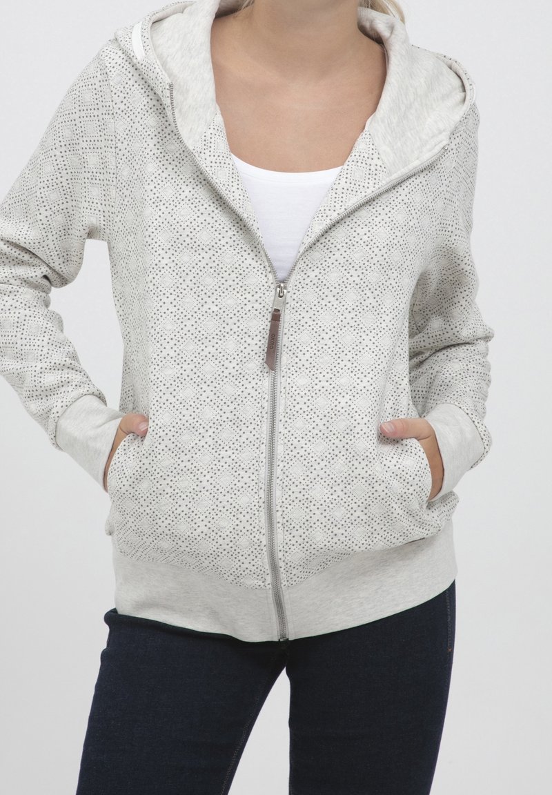 Femme portant un sweat à capuche à motif gris clair avec zip, mains dans les poches, associé à un jean bleu foncé et un t-shirt blanc en dessous.