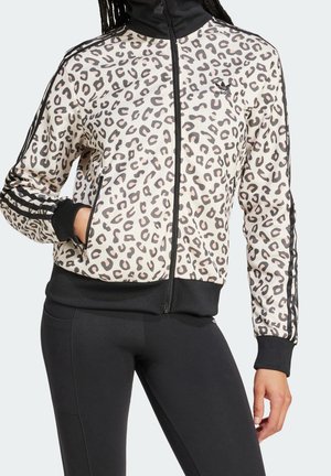 Femme portant une veste zippée Adidas blanche et noire à imprimé léopard avec des poignets noirs et des bandes sur les manches, et un pantalon noir.
