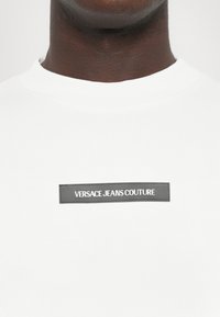 T-shirt bianco in cotone con colletto rotondo, caratterizzato da una patch rettangolare nera con la scritta "VERSACE JEANS COUTURE" in bianco.