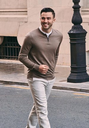 Smiling man wearing brown zip-neck sweater and light checkered pants walking on city street near black lamp post and stone building.