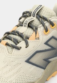 Pantof sport bej cu material din plasă, șireturi gri, accente portocalii și talpă de cauciuc texturată. Prezintă un logo proeminent pe lateral.