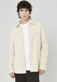 Chemise à boutons couleur crème avec un design texturé en weave gaufré, manches longues et poche poitrine. Portée sur un t-shirt blanc.