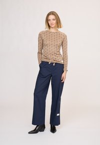 Beżowy top z długim rękawem z powtarzającym się wzorem wstążki, w połączeniu z granatowymi spodniami o szerokich nogawkach i czarnymi botkami. Gładkie tekstury na całej powierzchni.