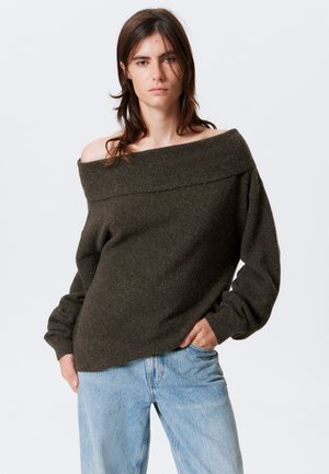 Olive green off-shoulder sweater with a wide ribbed neckline, textured fabric, and loose-fit sleeves, paired with light blue jeans.