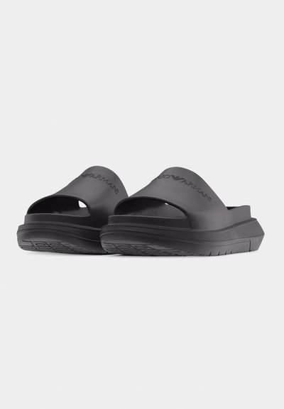 Μαύρα slides slip-on με ανυψωμένες σόλες και ανάγλυφο λογότυπο "Emporio Armani" στα λουράκια, τοποθετημένα σε γωνία πάνω σε απλό φωτεινό φόντο.