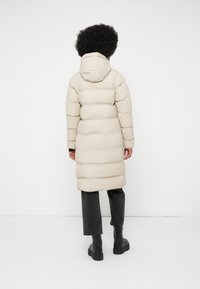 Beige pufferjakke med vertikalt quiltet design, hætte og lang pasform. Sammen med mørke bukser og kraftige sorte støvler.