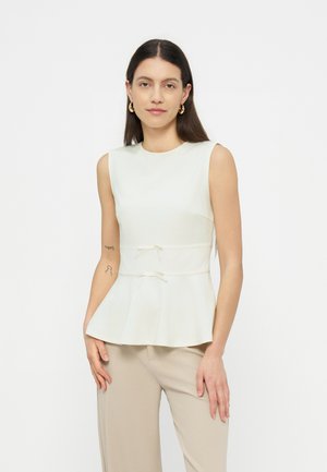 Kvinne med langt mørkt hår iført hvit ermeløs peplum-topp og beige bukser, stående mot en ensfarget hvit bakgrunn.