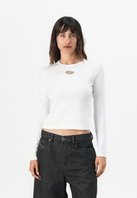 Haut blanc à manches longues avec encolure ronde et accent logo en métal, associé à un denim foncé. Le tissu est lisse avec une silhouette ajustée.