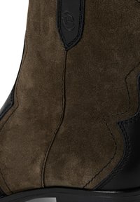 Braune Wildleder-Stiefelette mit schwarzen Lederakzenten und dekorativen Nähten. Die geschwungene Form und die glatte Textur betonen die Verarbeitung.