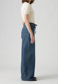 Ljusbeige kortärmad topp parat med högmidjade, vida mörkblå jeans med knytmidja och bakfickor.