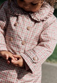 Manteau matelassé à carreaux rose et crème avec un col à volants et des accents de boutons. Détails visibles aux poignets et mains jointes devant.