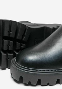 Botas de cuero negro con punta redondeada, suela gruesa y estriada, y detalles cosidos a lo largo del borde. La suela exterior presenta patrones de tracción profundos.