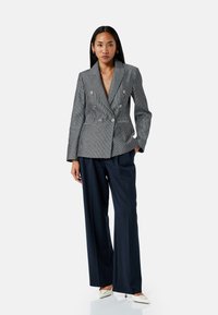 Femme se tenant debout avec une main dans la poche, portant un blazer gris à rayures et double boutonnage, un pantalon large bleu marine et des talons aiguilles blancs.