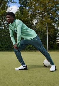 Mintgrön långärmad skjorta med texturerade ränder, i kombination med mörkgröna smala byxor. Vita och svarta fotbollsskor. Fotboll i närheten.