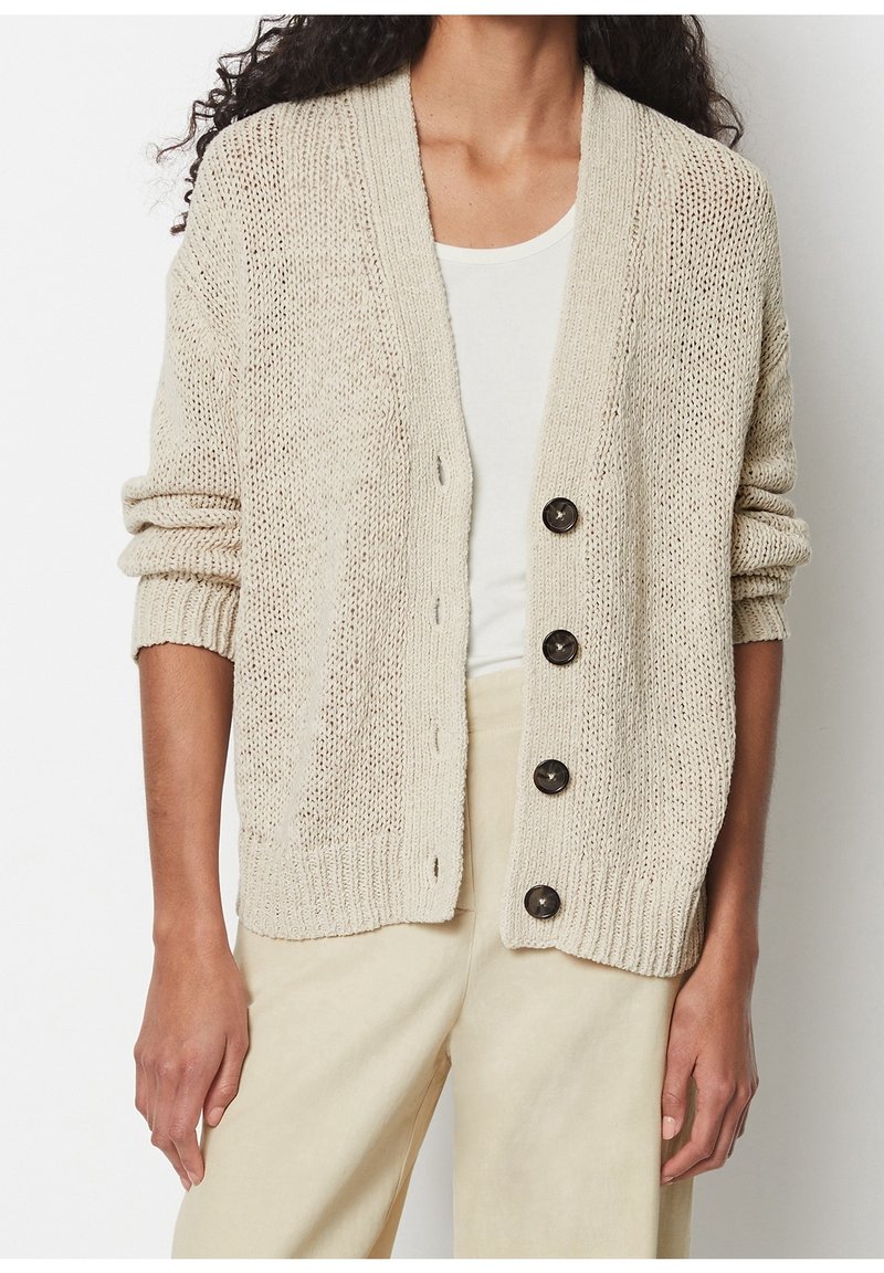 Personne portant un cardigan en maille beige avec quatre boutons foncés sur un haut blanc et un pantalon beige clair, les bras détendus le long du corps.