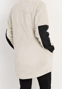 Fleece-Mantel mit einem cremefarbenen, strukturierten Oberteil und schwarzen, glatten Ärmel. Mit abgerundetem Saum und seitlichen Taschen.
