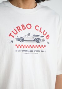 Balta marškinėliai su grafika, vaizduojančia kabrioletą. Raudonai ir mėlynai parašyta "TURBO KLUBAS" ir "AUKŠTOS KOKYBĖS SPORTINIAI AUTOMOBILIAI."