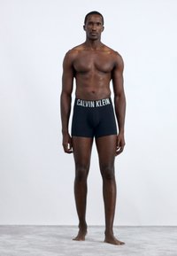 Fekete pamut boxer alsónadrág széles derékpánttal, rajta fehér "CALVIN KLEIN" felirattal. A modell mezítláb áll, előre nézve, egy világos háttér előtt.