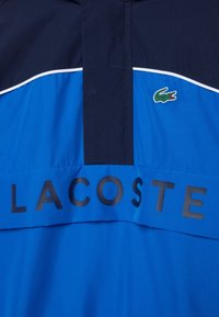 Blue and navy jacket with white piping, black zipper flap, Lacoste text across front, and small green crocodile logo on right chest.