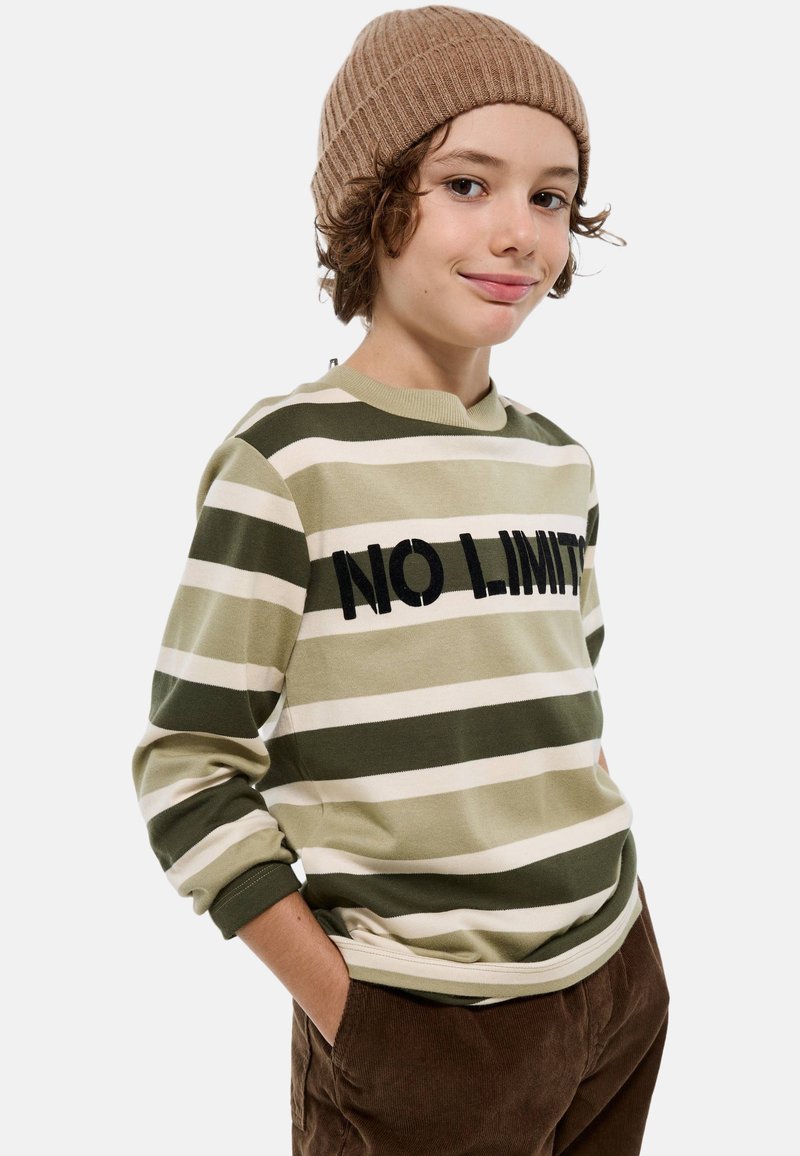 Sweatshirt rayé vert et crème avec "NO LIMITS" en texte noir, associé à un pantalon en velours côtelé marron. Modèle portant un bonnet en tricot beige.