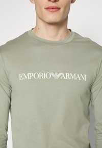 Nahaufnahme einer Person, die ein hellgrünes Langarmshirt mit "EMPORIO ARMANI" und dem weißen Markenlogo des Adlers auf der Brust trägt.