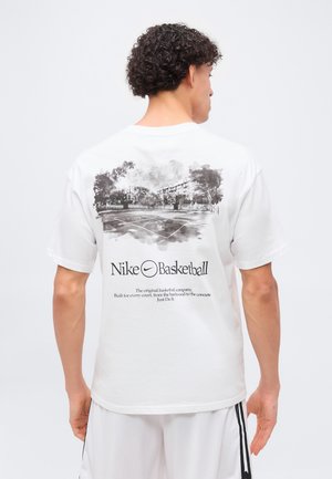 Mężczyzna zwrócony tyłem nosi białą koszulkę Nike Basketball z czarnym szkicem zewnętrznego boiska do koszykówki i tekstem poniżej na jednolitym tle.