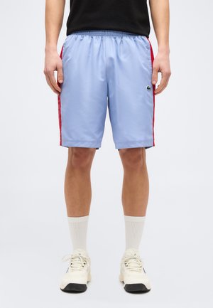 Person wearing light blue athletic shorts with red side stripes, white crew socks, and white sneakers, standing against a plain white background.