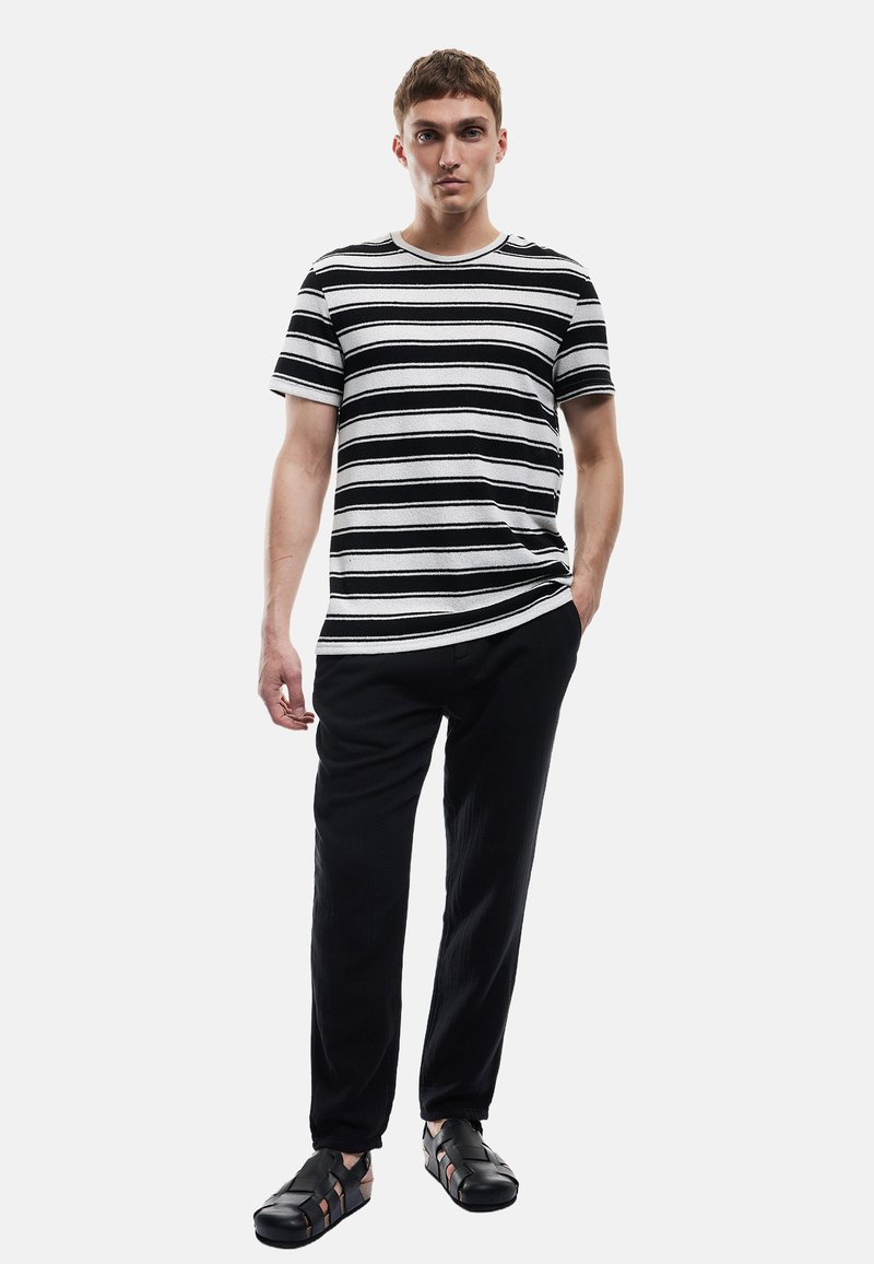 Sort og hvid stribet T-shirt med korte ærmer, parret med sorte bukser og sorte sandaler med stropper. Enkel design, glat stoftekstur.