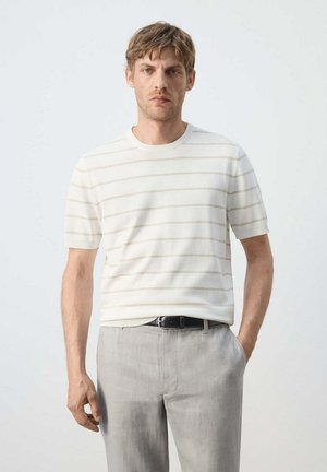 Hombre de pie con una mano en el bolsillo, vestido con camisa blanca de manga corta con finas rayas horizontales y pantalones gris claro con cinturón negro.