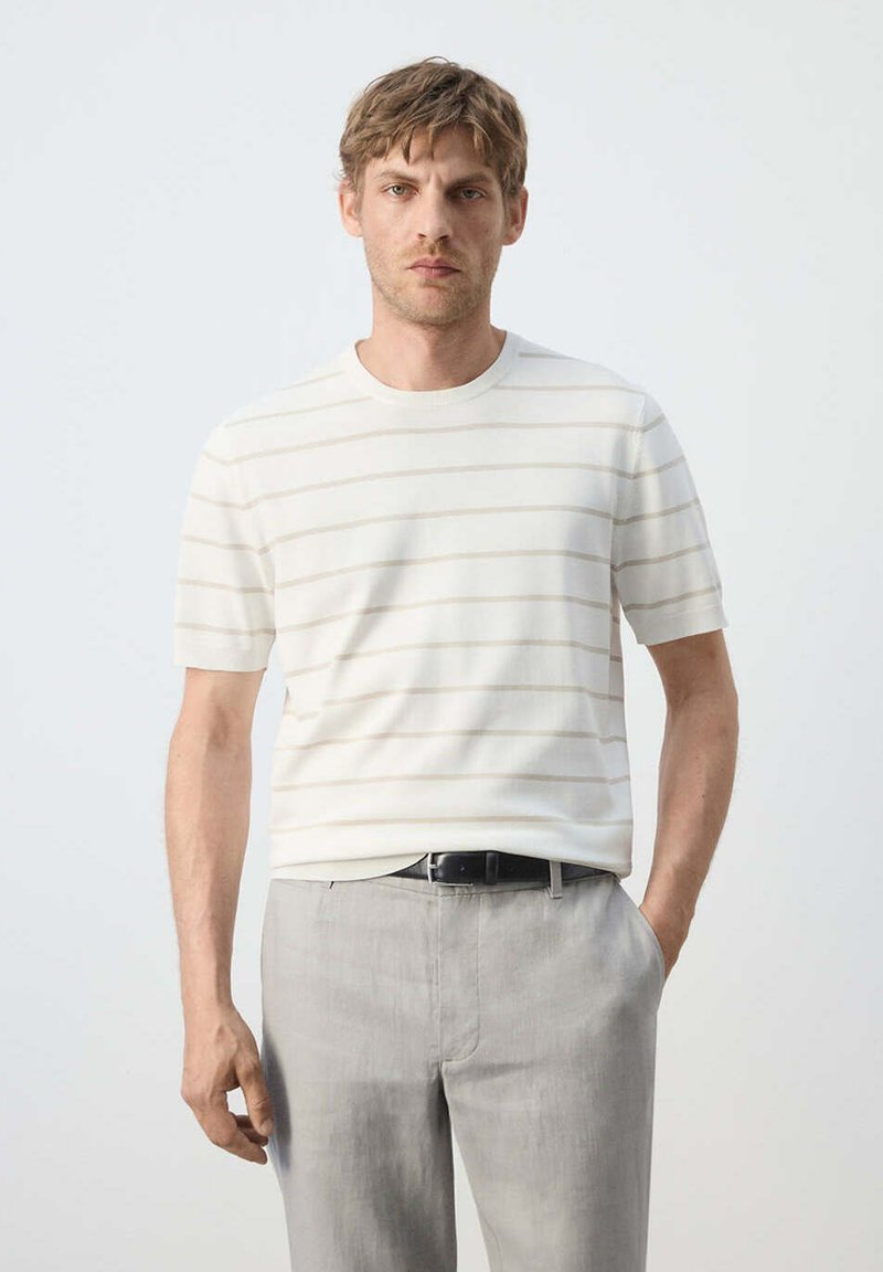 Homme debout, une main dans la poche, portant une chemise blanche à manches courtes avec de fines rayures horizontales et un pantalon gris clair avec une ceinture noire.