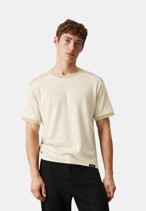 Giovane uomo con capelli ricci che indossa una maglietta a maniche corte a coste color crema con bordi beige e pantaloni neri, in piedi davanti a uno sfondo semplice.