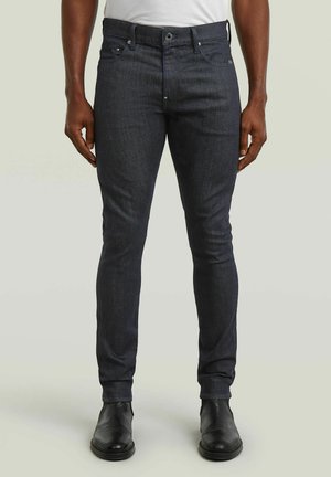 Mann trägt eng anliegende dunkelgraue Jeans, weißes Hemd und schwarze Lederstiefel, steht vor einem schlichten hellen Hintergrund.