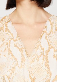 Gros plan sur le cou et le haut de la poitrine d'une personne portant un vêtement à motif serpent beige et blanc en V, avec une coupe de cheveux brun foncé courte visible.